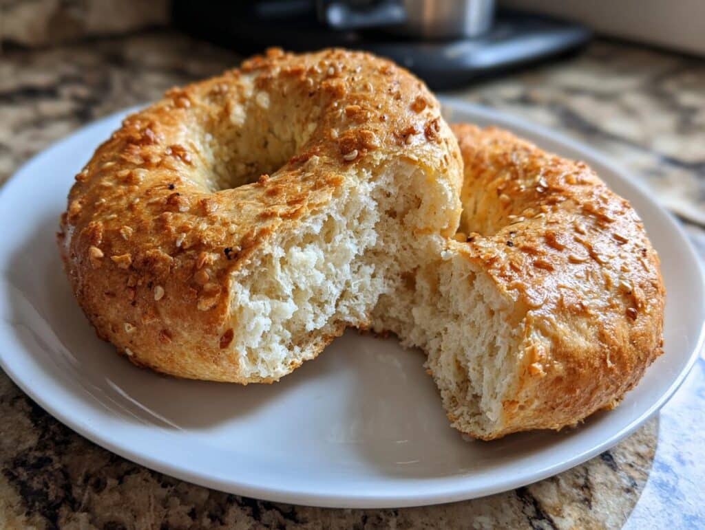 Two homemade protein bagels on a white plate, one bagel torn in half to show the inside texture.