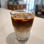 A glass of Iced Caramel Macchiato with layers of milk, coffee, caramel and ice on a white surface.