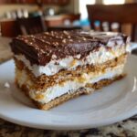 A slice of No-Bake Eclair Cake on a white plate, showing layers of graham crackers, cream, and chocolate frosting.