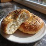 An everything bagel, cut in half and displayed on a white plate. The bagel is topped with sesame and poppy seeds.
