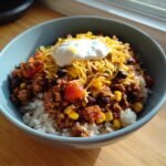 A bowl of rice topped with seasoned ground meat, corn, black beans, tomatoes, cheese, and sour cream. High-protein meal.