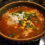 A bowl of Mexican chicken tortilla soup with rice, garnished with corn, tomatoes, and cilantro.