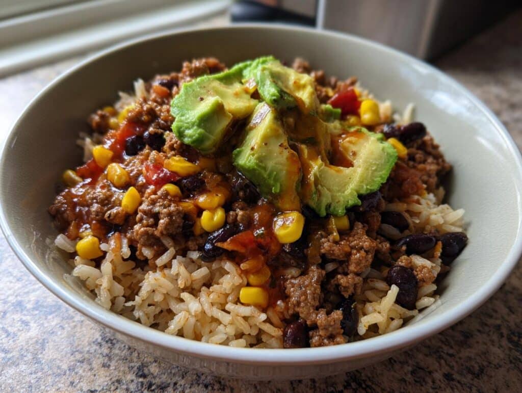 A delicious Mexican rice bowl with sauce, ground beef, corn, black beans, and fresh avocado slices.