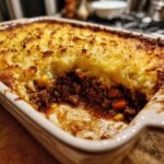 Close-up of a partially eaten shepherds pie casserole in a white baking dish, showing the meat and potato layers.