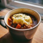 A mug of shepherds pie soup recipe easy, topped with mashed potatoes and herbs, sitting on a wooden surface.